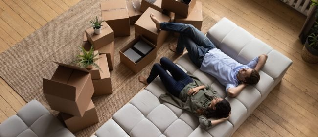 Top view happy couple tenants homeowners relaxing on couch on moving day in living room with cardboard boxes, man and woman resting after relocation in first own apartment, mortgage or rent